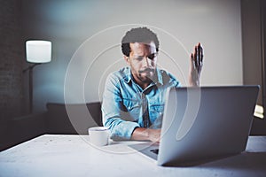 Indignant bearded African man making video conversation via laptop with partners while working at coworking office