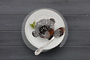 An individual chocolate sponge cake, with a sticky chocolate sauce, on a white plate with a spoon, shot on a dark grey wooden