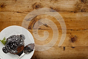 An individual chocolate sponge cake, with a sticky chocolate sauce, on a white plate with a spoon, shot on a wooden background