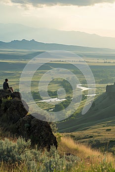 Individual sitting in solitude on a hill, overlooking a tranquil valley with a river snaking through it at dusk. photo