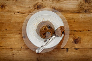 An individual spotted dick sponge cake, on a white plate with a spoon and sprinckled brown sugar, on a wooden surface