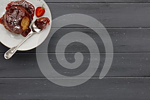 An individual strawberry sponge cake, with a sticky strawberry sauce, on a white plate with a spoon, on a dark grey wooden surface