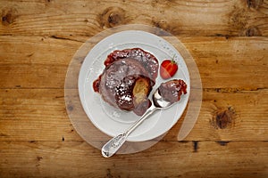 An individual strawberry sponge cake, with a sticky strawberry sauce, on a white plate with a spoon, on a wooden surface with