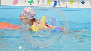 Individual swimming lesson with trainer in an indoor pool. Happy preschool girl is learning to swim with pool noodle in