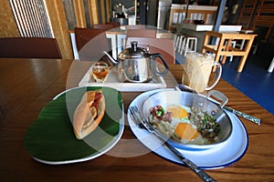 Indochina pan-fried egg with toppings with Baguette bread sandwich with cheese, ham on fresh Green banana leaf and ice coffee, tea