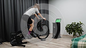 Indoor Cycling Concept. Young female cyclist pedaling bicycle on smart trainer looking at computer screen.