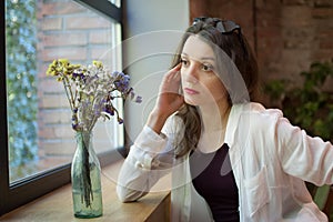 Indoor female portrait of a thoughtful girl in black top and white shirt sitting near the window in loft style cafe