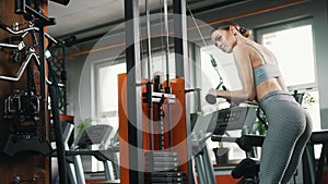 Indoor gym shot of a skinny caucasian young adult woman with slicked back hair trying out professional gym equipment for
