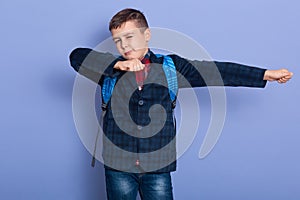 Indoor image of adorable playful little boy raising arms, closing one eye, wearing jeans, plaid shirt and jacket, studying hard,