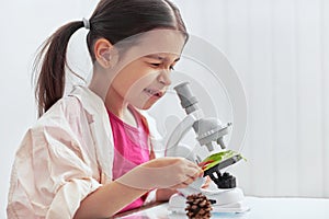 Indoor image of cute little girl working with microscope and exploring plants. The kid in lab coat learning science in the school