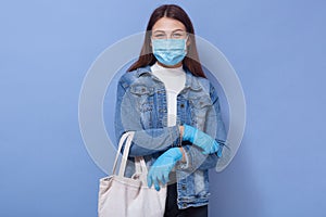 Indoor image of protected young female with long hair holding ecobag looking directly at camera, wearing antibacterial mask and