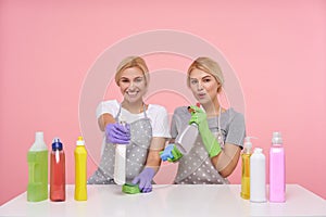 Indoor photo of lovely young white-headed housewives being in nice mood while making spring cleaning, sitting over pink background