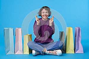 Indoor photo of young dark haired curly woman dressed in purple sweatshirt, jeans and sneakers closing one eye and crossing