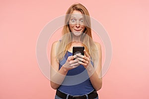 Indoor photo of young lovely redhead woman with casual hairstyle keeping mobile phone in raised hands and being surprised while