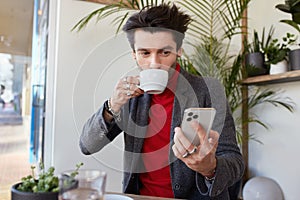 Indoor photo of young pretty brown haired male dressed in elegant clothes while having cup of coffee in city cafe, keeping