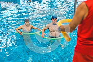 Indoor pool fitness exercises. Adult caucasian seniors doing aerobics exercises with foam pool noodles. Unrecognizable