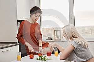 Indoor portrait of attractive hot woman sitting on table cutting tomotoe while her girlfriend drinks juice in kitchen