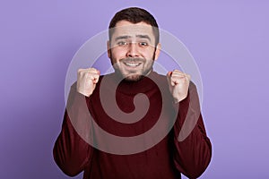 Indoor portrait of emotional charismatic good looking man with beard clenching fists, making gesture, smiling sincerely, having photo