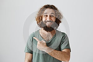 Indoor portrait of handsome kind arabian man with curly hair and beard smiling broadly while pointing left or behind