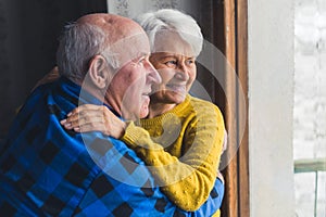 Indoor portrait of a happy caucasian heterosexual retired couple of seniors standing by the window embracing each other.