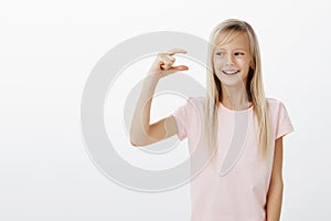 Indoor portrait of happy satisfied adorable young girl with fair hair, looking at tiny thing, shaping it with fingers