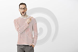 Indoor portrait of pleased impressed handsome man in eyewear, pointing at upper right corner, dropping jaw from positive