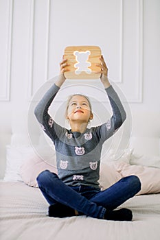 Indoor portrait of pretty school girl in casual domestic wear, sitting on the bed in lotus position and holding stylish