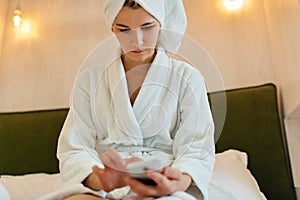 Indoor portrait of serious woman sitting on the bed after bath shower with towel texting messages on mobile phone. Beauty spa at