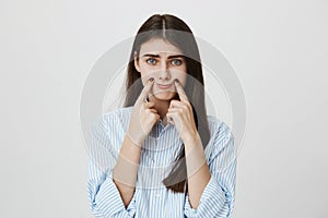 Indoor portrait of unhappy and stressed woman with frowned eyebrows stretching mouth with index fingers to make gloomy