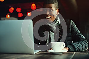 Indoor portrait of young black man sitting in cafe, drinking coffee or tea and working with lap top. Model looking at
