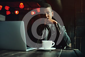 Indoor portrait of young black man sitting in cafe, drinking coffee or tea and working with lap top. Model looking at