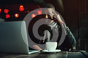 Indoor portrait of young black man sitting in cafe, drinking coffee or tea and working on lap top. Model looking at