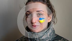 Indoor portrait of young girl with blue and yellow ukrainian flag on her cheek wearing military uniform, mandatory