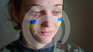 Indoor portrait of young girl with blue and yellow ukrainian flag on her cheek wearing military uniform, mandatory