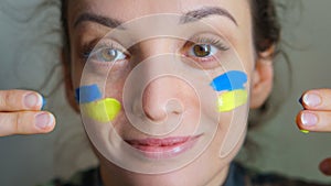 Indoor portrait of young girl with blue and yellow ukrainian flag on her cheek wearing military uniform, mandatory