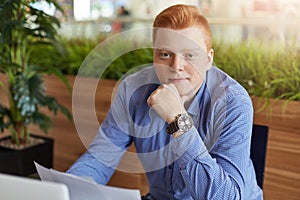 Indoor portrait of young stylish businessman with red hair dressed in checked shirt sitting a office working trough papers, studyi