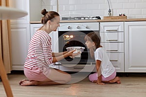 Indoor profile shot of smiling taking baking out of oven, her daughter standing near by and smelling delicious sweets, kid heping