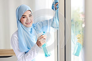 Indoor shot active young beautiful asian muslim housewife woman holding cleaning spray, washing window glass. Portrait cute girl w