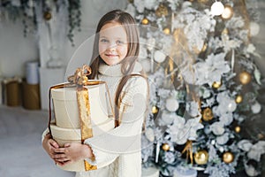 Indoor shot of adorable cute pretty small female child wears warm knitted clothes, holds huge wrapped present gift, recieves from