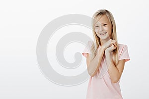 Indoor shot of adorable joyful young female child with blond hair, holding hands together near face and smiling broadly