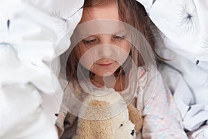 Indoor shot of adorable little child girl playing with teddy bear in bed under blanket, charming kid plays in morning before