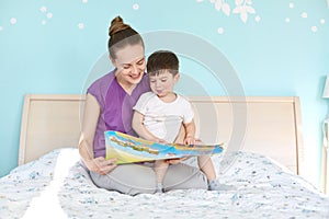 Indoor shot of affectionate woman and small boy read interesting story from book, sit at bed, enjoy calm atmosphere, smile
