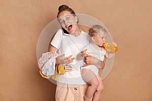 Indoor shot of amazed excited joyful mother with her baby standing and talking on mobile phone, hearing good news, spending