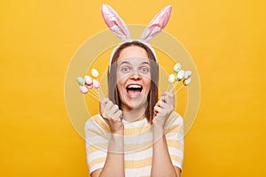 Indoor shot of amazed overjoyed woman dressed rabbit ears holding Easter cake pops , screaming with happiness, celebrating with