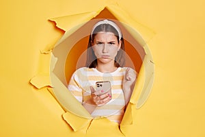 Indoor shot of angry upset woman wearing striped t shirt looking through breakthrough of yellow background, holding mobile phone