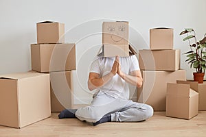Indoor shot of anonymous man wearing white T-shirt sitting on the floor near cardboard boxes, posing with carton box on his head