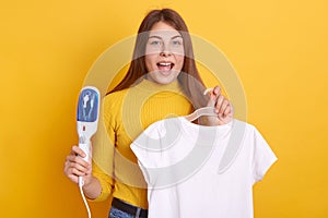Indoor shot of astonished woman wearing casual clothing holding curling iron and white t shirt on hangers, girl going on date,