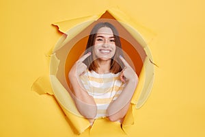 Indoor shot of attractive adorable smiling young woman wearing striped T-shirt, pointing at her white perfect teeth, breaking