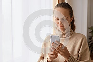 Indoor shot of attractive smiling female woman wearing beige sweater standing near window with cell phone in hands, scrolling