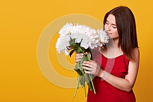 Indoor shot of attractive tender sweet lady in red dress posing isolated over yellow background, holding white flowers in both